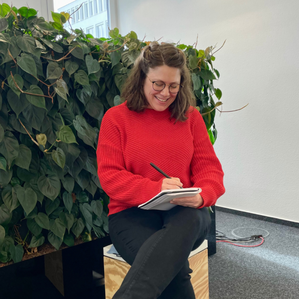 Teamleiterin Pia Wiedenbruch sitzt im Büro auf einem Hocker vor einer Pflanzwand und schreib mit einem Stift in ihr Notizbuch.